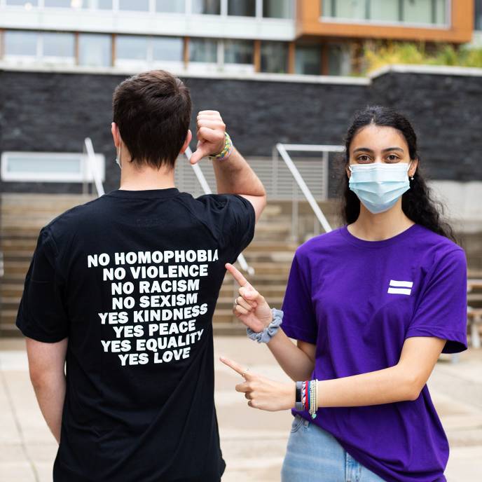 One student pointing to the shirt of another. Shirt says 'No Homophobia, No Violence, No Racism, etc.'